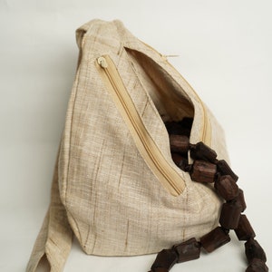 K&ouml;nnte beinhalten: Eine beige Leinen-Tasche mit einem Rei&szlig;verschlussfach und einer braunen Holzperlenkette im Inneren. Die Tasche hat einen langen Riemen und eine kleine Schlaufe an der Seite. Die Tasche ist mit "Mayapur Store" beschriftet.