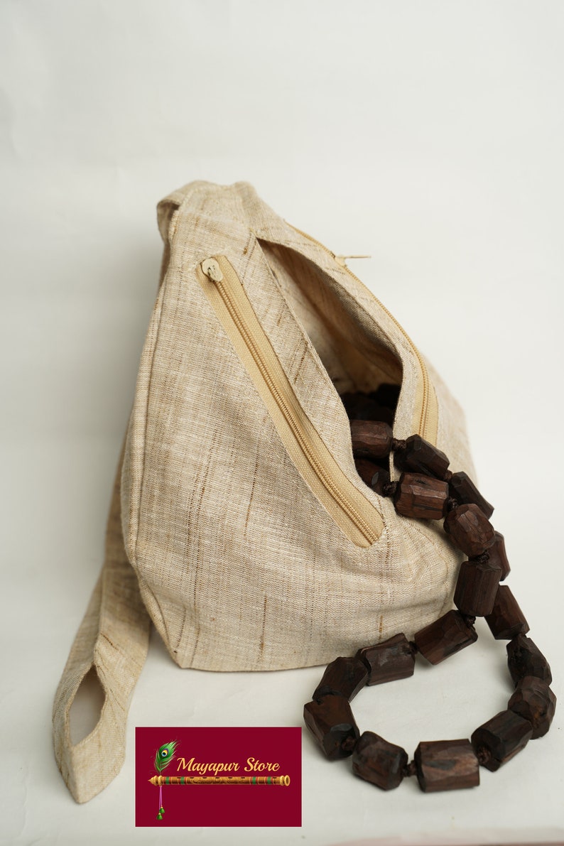 K&ouml;nnte beinhalten: Eine beige Leinen-Tasche mit einem Rei&szlig;verschlussfach und einer braunen Holzperlenkette im Inneren. Die Tasche hat einen langen Riemen und eine kleine Schlaufe an der Seite. Die Tasche ist mit "Mayapur Store" beschriftet.
