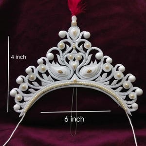 以下が含まれることがあります： パールアクセントとピンクの羽根が付いた白いティアラ。ティアラの高さは10cm、幅は15cmです。白鳥のような形と小さな真珠のデザインです。ティアラには頭に固定するためのワイヤーが付いています。