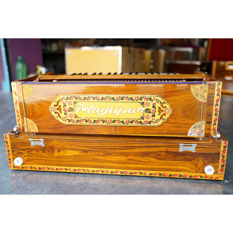 May include: A wooden harmonium with a polished finish. The instrument displays the word "Mayapur" in a decorative panel. The harmonium has a keyboard and is decorated with floral patterns in red, black, and gold.