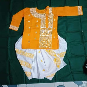 May include: An orange kurta and white dhoti set with yellow and white decorative patterns. The kurta has long sleeves and a detailed neckline. The dhoti is white with yellow accents. The outfit is displayed on a green surface.