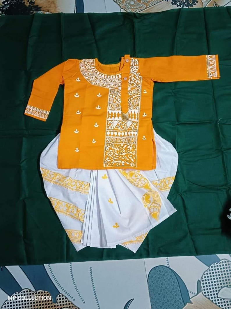 May include: An orange kurta and white dhoti set with yellow and white decorative patterns. The kurta has long sleeves and a detailed neckline. The dhoti is white with yellow accents. The outfit is displayed on a green surface.