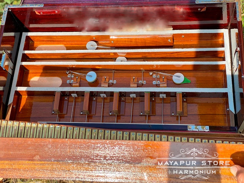 Pode incluir: Um harm&oacute;nio de madeira com acabamento castanho e interior branco. O harm&oacute;nio tem uma fila de teclas e um fole. O harm&oacute;nio est&aacute; aberto e o fole &eacute; vis&iacute;vel. O harm&oacute;nio est&aacute; etiquetado como "MAYAPUR STORE HARMONIUM".