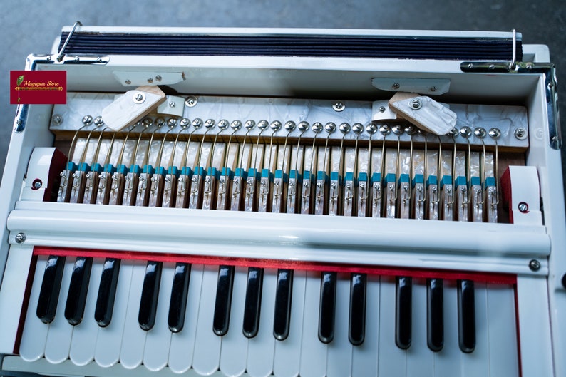 May include: A white harmonium with a red trim and a keyboard with black and white keys. The harmonium is open, revealing the internal workings of the instrument.