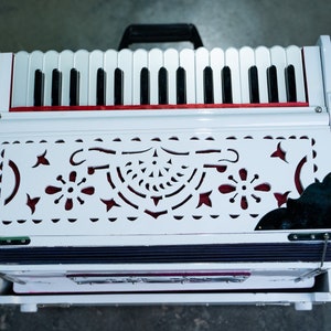 May include: A white harmonium with a red trim and a decorative, cut-out design on the front. The harmonium has a black and white keyboard and a bellows system.