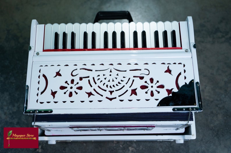 May include: A white harmonium with a red trim and a decorative, cut-out design on the front. The harmonium has a black and white keyboard and a bellows system.