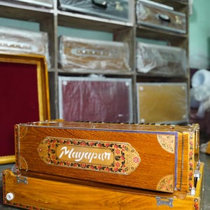 K&ouml;nnte beinhalten: Ein Harmonium aus Holz mit aufwendigen floralen Verzierungen und dem Wort "Mayapur" auf der Vorderseite. Das Harmonium befindet sich in einem Gesch&auml;ft mit anderen Musikinstrumenten in Regalen im Hintergrund.