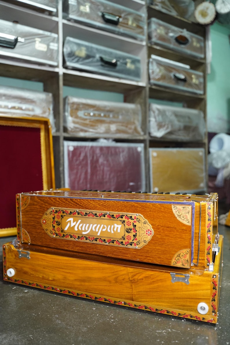 K&ouml;nnte beinhalten: Ein Harmonium aus Holz mit aufwendigen floralen Verzierungen und dem Wort "Mayapur" auf der Vorderseite. Das Harmonium befindet sich in einem Gesch&auml;ft mit anderen Musikinstrumenten in Regalen im Hintergrund.