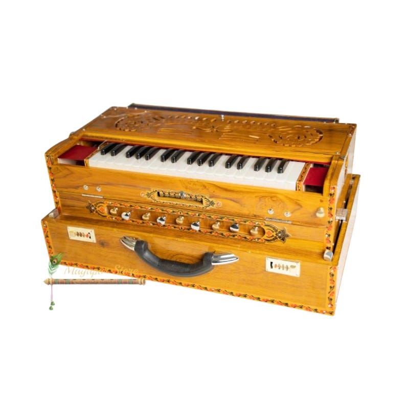 May include: A wooden harmonium with a keyboard, buttons, and a handle. The instrument is light brown with decorative carvings and painted accents. The keyboard has white and black keys. A flute is visible in the lower left corner.