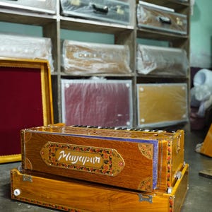 K&ouml;nnte beinhalten: Ein Harmonium aus Holz mit aufwendigen floralen Verzierungen und dem Wort "Mayapur" auf der Vorderseite. Das Harmonium ist braun mit goldenen Akzenten und steht auf einem Betonboden.