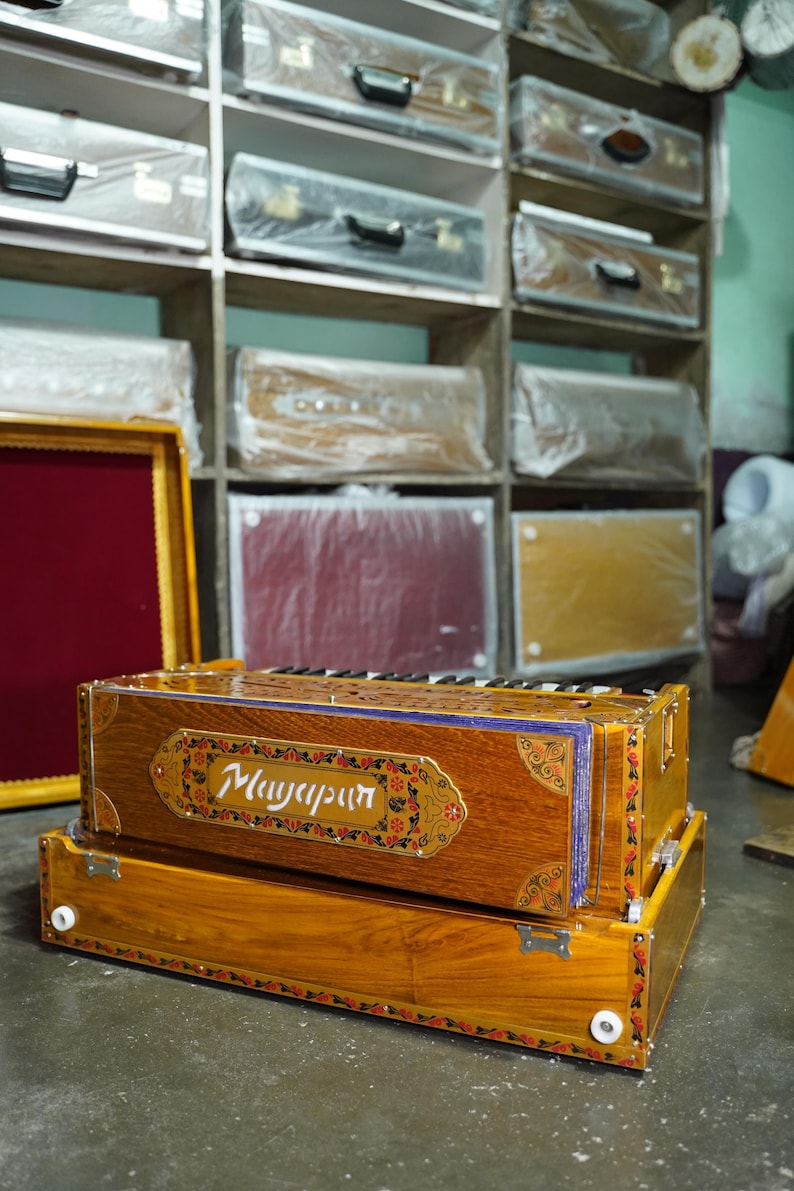 K&ouml;nnte beinhalten: Ein Harmonium aus Holz mit aufwendigen floralen Verzierungen und dem Wort "Mayapur" auf der Vorderseite. Das Harmonium ist braun mit goldenen Akzenten und steht auf einem Betonboden.