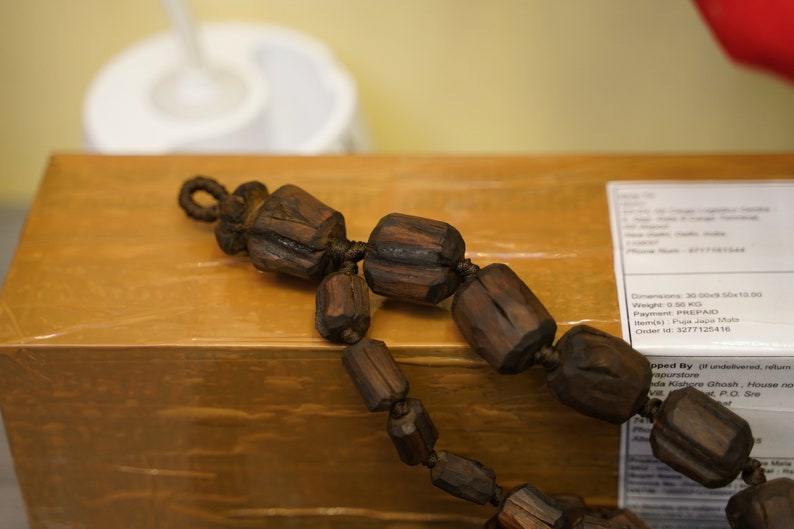 K&ouml;nnte beinhalten: Eine braune Holzperlenkette mit einer geknoteten Schnur, die die Perlen verbindet. Die Kette liegt auf einem braunen Karton mit einem Versandetikett.