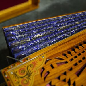 K&ouml;nnte beinhalten: Ein Holzmusikinstrument mit einem blau und gold lackierten Blasebalg und aufwendigen Schnitzereien. Das Instrument hat eine Reihe von L&ouml;chern auf der Vorderseite und ein dekoratives Blumenmuster.