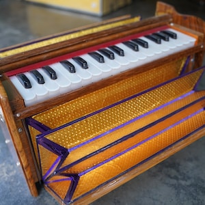 May include: A wooden harmonium with a keyboard of black and white keys. The instrument has a gold and purple patterned bellows and a red accent above the keys. The harmonium is a traditional Indian instrument.