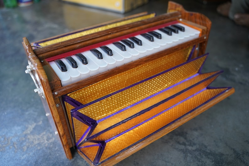 May include: A wooden harmonium with a keyboard of black and white keys. The instrument has a gold and purple patterned bellows and a red accent above the keys. The harmonium is a traditional Indian instrument.
