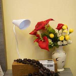 K&ouml;nnte beinhalten: Eine wei&szlig;e Schreibtischlampe mit flexiblem Arm und wei&szlig;em Schirm steht auf einem Tisch neben einem braunen Karton. Ein Strau&szlig; roter und gelber Blumen in einer wei&szlig;en Vase steht auf dem Tisch. Eine lange Schnur aus braunen Holzperlen liegt auf dem Tisch.