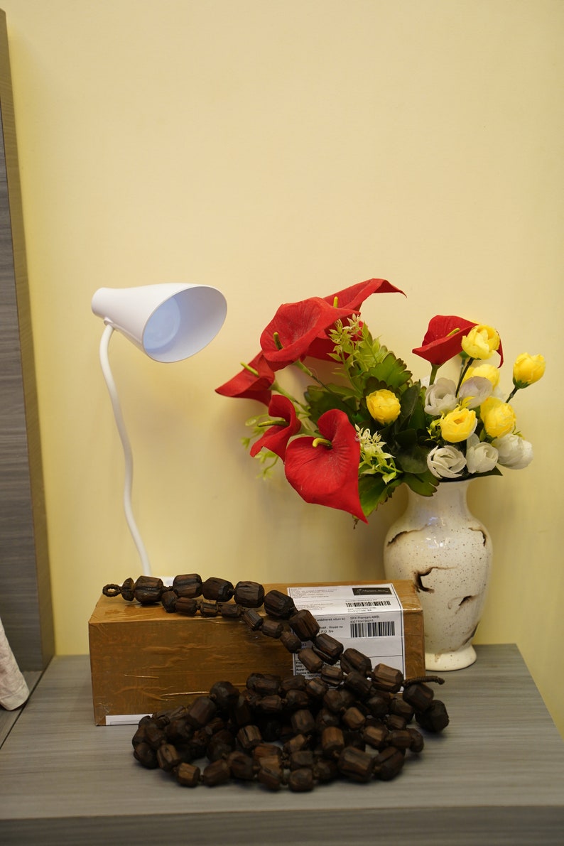 K&ouml;nnte beinhalten: Eine wei&szlig;e Schreibtischlampe mit flexiblem Arm und wei&szlig;em Schirm steht auf einem Tisch neben einem braunen Karton. Ein Strau&szlig; roter und gelber Blumen in einer wei&szlig;en Vase steht auf dem Tisch. Eine lange Schnur aus braunen Holzperlen liegt auf dem Tisch.