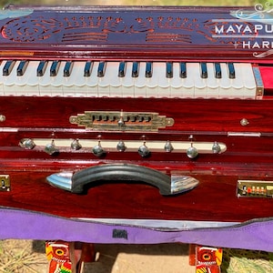 Pode incluir: Um harm&oacute;nio de madeira vermelho escuro com um teclado branco e uma pega preta. O harm&oacute;nio tem um topo de madeira esculpida decorativo com o texto "MAYAPUR STORE HARMONIUM".