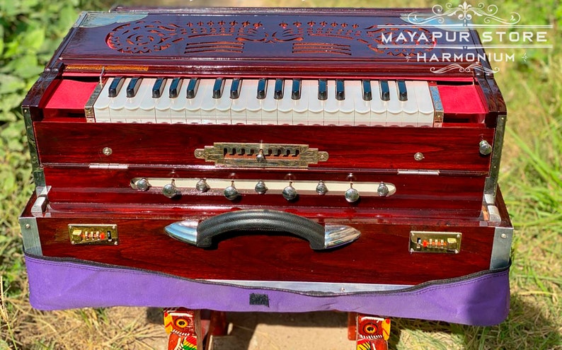 Pode incluir: Um harm&oacute;nio de madeira vermelho escuro com um teclado branco e uma pega preta. O harm&oacute;nio tem um topo de madeira esculpida decorativo com o texto "MAYAPUR STORE HARMONIUM".