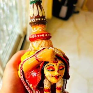 May include: A hand-painted ceramic figurine of a woman in a red and yellow sari, with a white floral design. The woman is wearing a traditional Indian bridal headdress and jewelry.