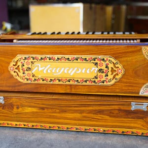 K&ouml;nnte beinhalten: Ein Harmonium aus Holz mit aufwendigen floralen Verzierungen und dem Wort "Mayurpur" auf der Vorderseite. Das Harmonium hat eine braune Holzoptik und einen dekorativen Rand.