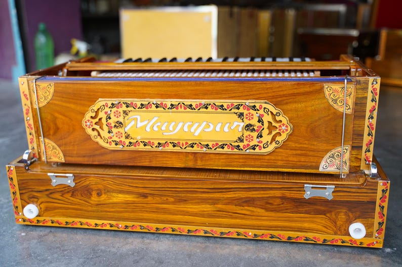 K&ouml;nnte beinhalten: Ein Harmonium aus Holz mit aufwendigen floralen Verzierungen und dem Wort "Mayurpur" auf der Vorderseite. Das Harmonium hat eine braune Holzoptik und einen dekorativen Rand.