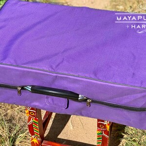 Pode incluir: Estojo de harm&oacute;nio roxo com al&ccedil;a preta e z&iacute;per. O estojo est&aacute; em um suporte de madeira com desenhos coloridos. O texto "MAYAPUR STORE HARMONIUM" &eacute; vis&iacute;vel no estojo.