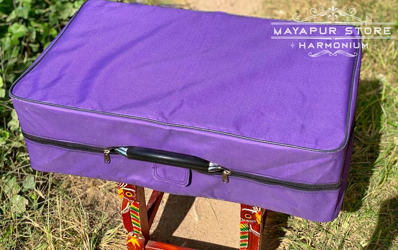 Pode incluir: Estojo de harm&oacute;nio roxo com al&ccedil;a preta e z&iacute;per. O estojo est&aacute; em um suporte de madeira com desenhos coloridos. O texto "MAYAPUR STORE HARMONIUM" &eacute; vis&iacute;vel no estojo.