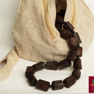 K&ouml;nnte beinhalten: Eine beigefarbene Leinentasche mit Rei&szlig;verschluss und einer braunen Holzperlenkette. Die Tasche hat einen Henkel und ist aus einem Naturstoff gefertigt.