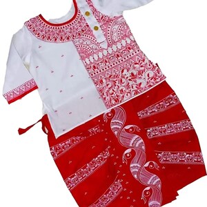 May include: A white and red traditional outfit. The top is white with red patterned accents and gold buttons. The skirt is red with a white peacock design and decorative borders. The outfit is likely for a cultural event or celebration.