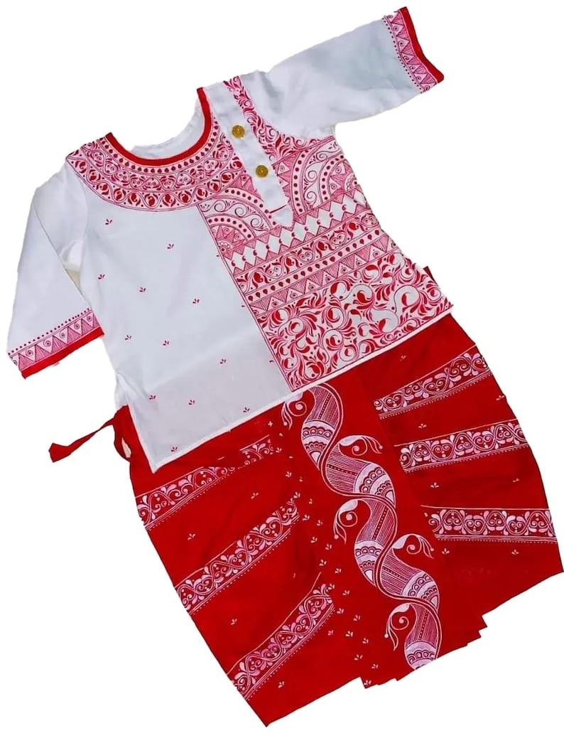 May include: A white and red traditional outfit. The top is white with red patterned accents and gold buttons. The skirt is red with a white peacock design and decorative borders. The outfit is likely for a cultural event or celebration.