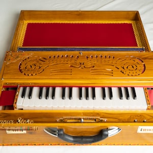 May include: A wooden harmonium with a keyboard, featuring intricate carvings and a red velvet interior. The instrument has a handle and is open, revealing the keys and bellows. The exterior is a warm, light brown colour.