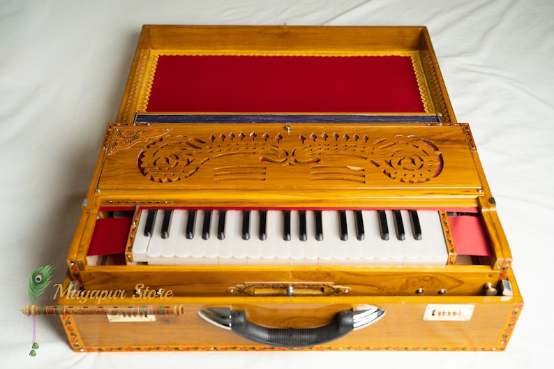 May include: A wooden harmonium with a keyboard, featuring intricate carvings and a red velvet interior. The instrument has a handle and is open, revealing the keys and bellows. The exterior is a warm, light brown colour.