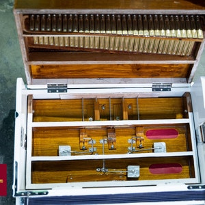 May include: An open wooden case with a white interior. The case has a row of metal reeds and a series of wooden levers and pedals. The case is open and the interior is visible. The case is made of wood and has a natural finish.