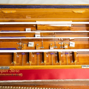 May include: A wooden harmonium with a polished finish, featuring internal mechanisms and a keyboard. The instrument has a red and purple fabric accent. The text "Mayapur Store" is visible on the lower left.