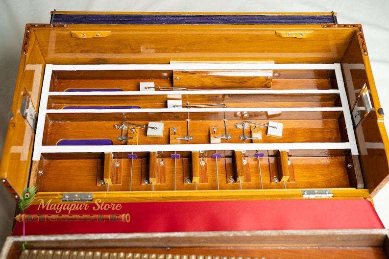 May include: A wooden harmonium with a polished finish, featuring internal mechanisms and a keyboard. The instrument has a red and purple fabric accent. The text "Mayapur Store" is visible on the lower left.