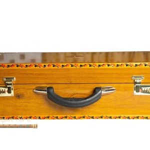 May include: A wooden suitcase with a black handle and two metal latches. The suitcase is a light brown colour with a decorative border of orange, black, and yellow floral designs. The suitcase has a combination lock on each side.