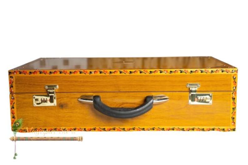 May include: A wooden suitcase with a black handle and two metal latches. The suitcase is a light brown colour with a decorative border of orange, black, and yellow floral designs. The suitcase has a combination lock on each side.