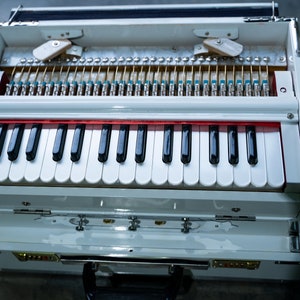 May include: A white harmonium with a red stripe above the keyboard. The harmonium has a wooden case with a hinged lid. The keyboard has 37 keys, with black and white keys.