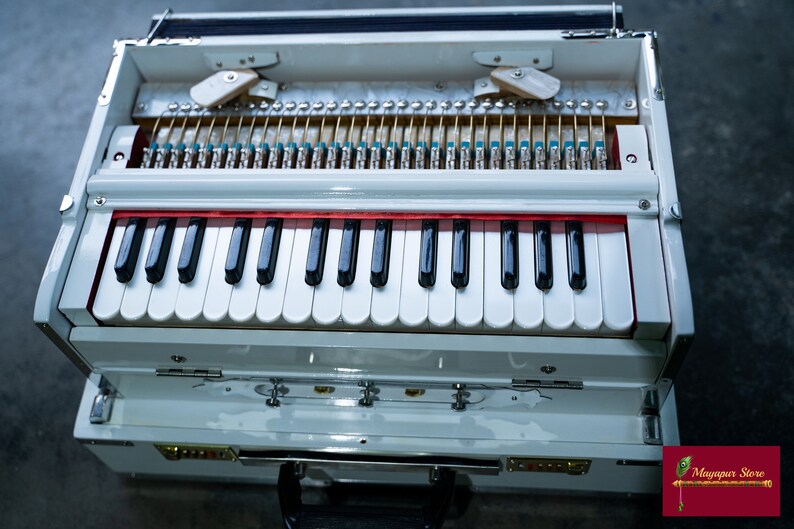 May include: A white harmonium with a red stripe above the keyboard. The harmonium has a wooden case with a hinged lid. The keyboard has 37 keys, with black and white keys.