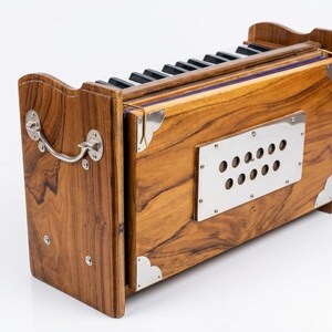 May include: A wooden harmonium with a natural wood grain finish. The instrument features a keyboard with black and white keys, a silver metal plate with holes, and silver corner accents. A silver handle is on the side.