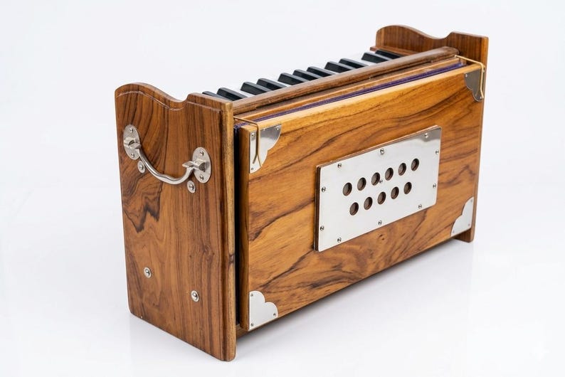 May include: A wooden harmonium with a natural wood grain finish. The instrument features a keyboard with black and white keys, a silver metal plate with holes, and silver corner accents. A silver handle is on the side.