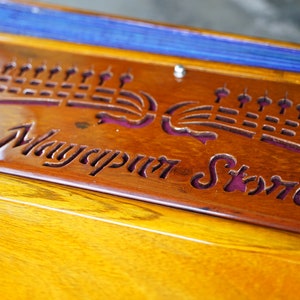 May include: Close-up of a wooden decorative piece with the text "Mayapur Store" carved into it. The wood has a warm, reddish-brown hue, with a glossy finish. The text is in a stylized font.