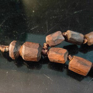 K&ouml;nnte beinhalten: Nahaufnahme einer Holzperlenkette. Die Perlen sind dunkelbraun, geometrisch und facettiert. Die Schnur hat eine passende braune Farbe. Die Halskette scheint handgefertigt zu sein und hat eine nat&uuml;rliche, rustikale &Auml;sthetik.