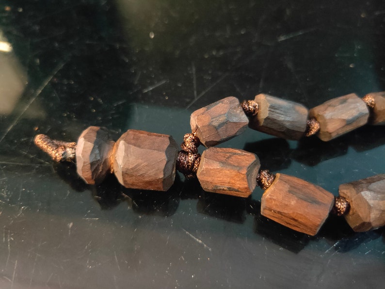K&ouml;nnte beinhalten: Nahaufnahme einer Holzperlenkette. Die Perlen sind dunkelbraun, geometrisch und facettiert. Die Schnur hat eine passende braune Farbe. Die Halskette scheint handgefertigt zu sein und hat eine nat&uuml;rliche, rustikale &Auml;sthetik.