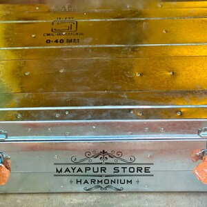 Pode incluir: Um ba&uacute; de armazenamento de metal prateado com duas travas e um r&oacute;tulo preto que diz "MAYAPUR STORE HARMONIUM".