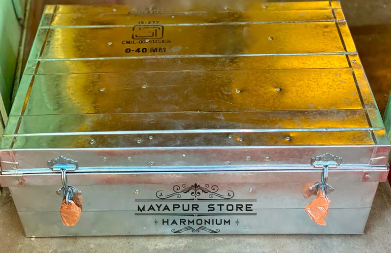 Pode incluir: Um ba&uacute; de armazenamento de metal prateado com duas travas e um r&oacute;tulo preto que diz "MAYAPUR STORE HARMONIUM".