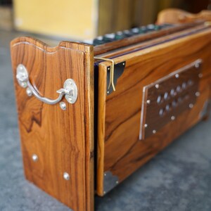 May include: A wooden harmonium with a rich, warm brown finish. The instrument features a silver handle and decorative metal accents. The design includes a rectangular metal plate with rivets. The harmonium is a traditional Indian musical instrument.