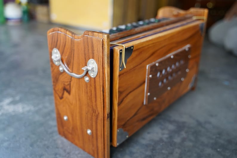 May include: A wooden harmonium with a rich, warm brown finish. The instrument features a silver handle and decorative metal accents. The design includes a rectangular metal plate with rivets. The harmonium is a traditional Indian musical instrument.