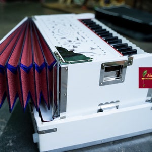 May include: A white harmonium with a red and blue bellows. The harmonium has a carved wooden design and a silver trim. The harmonium is open and the bellows are extended. The harmonium is sitting on a grey floor. Mayapur Store is printed on a red label.